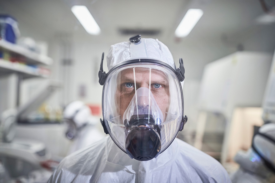 Man in PPE staring straight, from behind a glass window in a lab